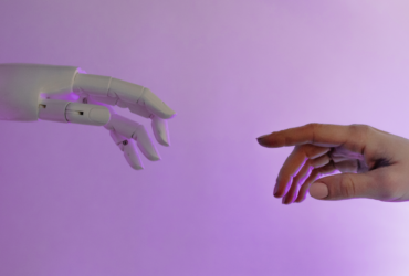 A robot hand on the left reaches out toward a human hand. They each have raised index fingers.