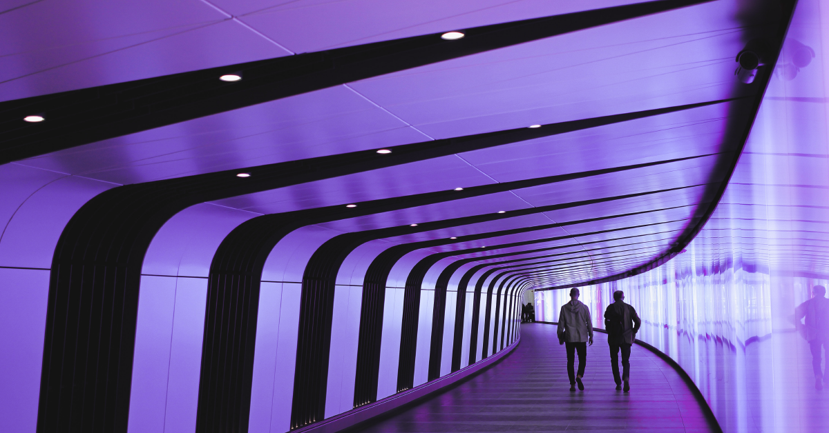 Two men walk away from the camera as they stroll through a modern tunnel with glass on one side and stripes on the other.